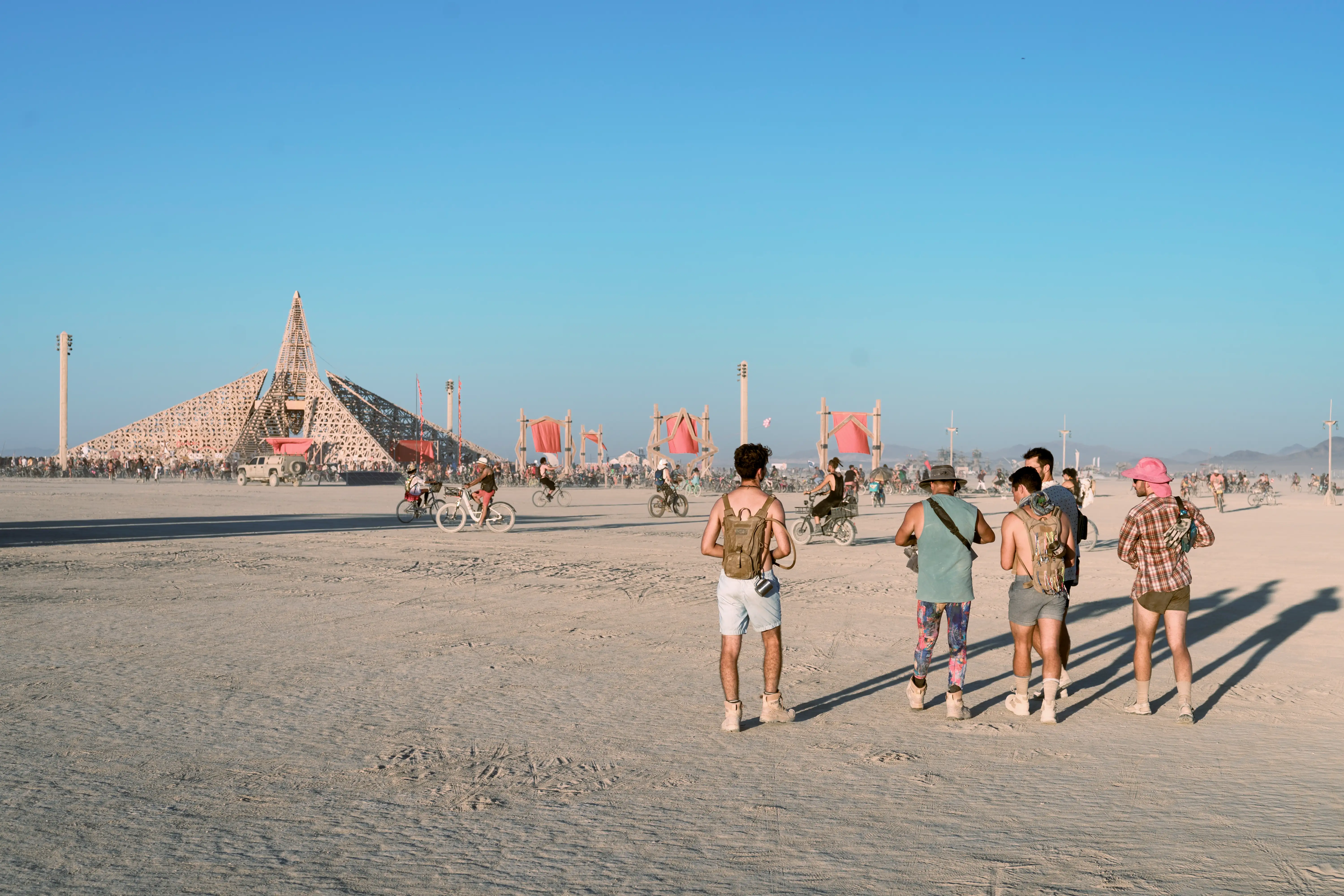 A group of five men, seen from behind, walking through the desert. They are approaching the Empyrean Temple on foot. It is a huge wood temple consisting of multiple angular shapes and patterns.